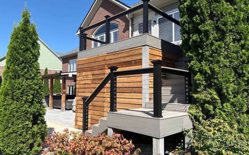 Modern backyard deck with wooden and composite materials, black metal railing, surrounded by lush greenery, perfect for outdoor entertaining and enjoying nature.